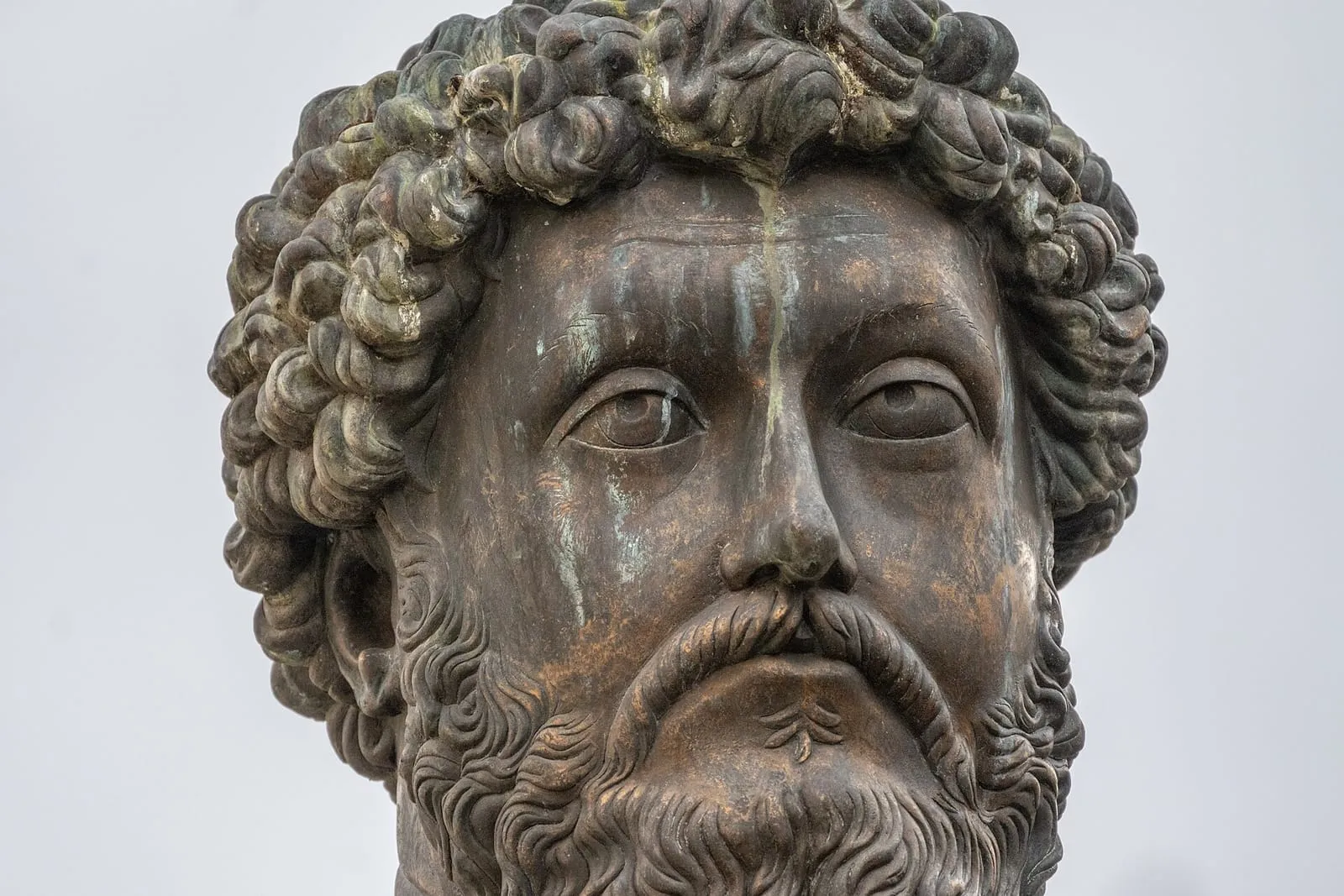 Rosto em bronze de Marco Aurélio, da estátua equestre do Capitólio, em Roma: cabelos e barba cacheados, olhar sereno, contra um céu claro.